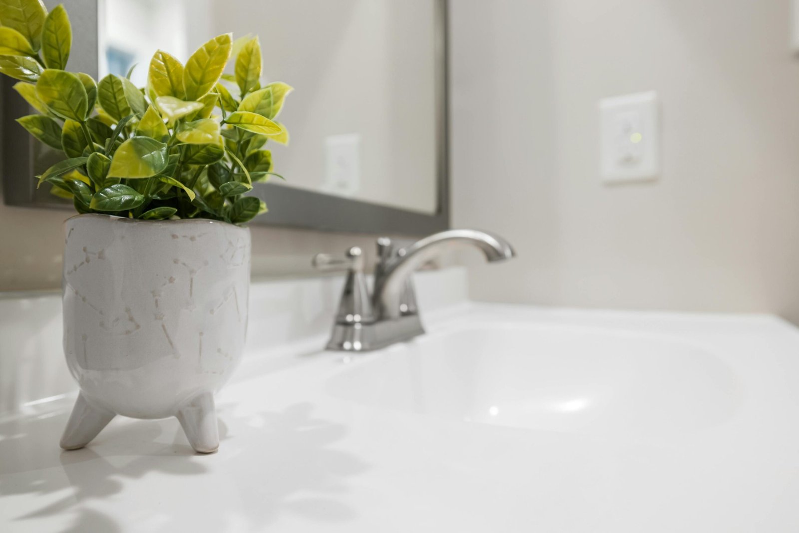 Planta verde em vaso de cerâmica branco com pés, posicionada sobre bancada de banheiro branca ao lado de uma torneira cromada e espelho.